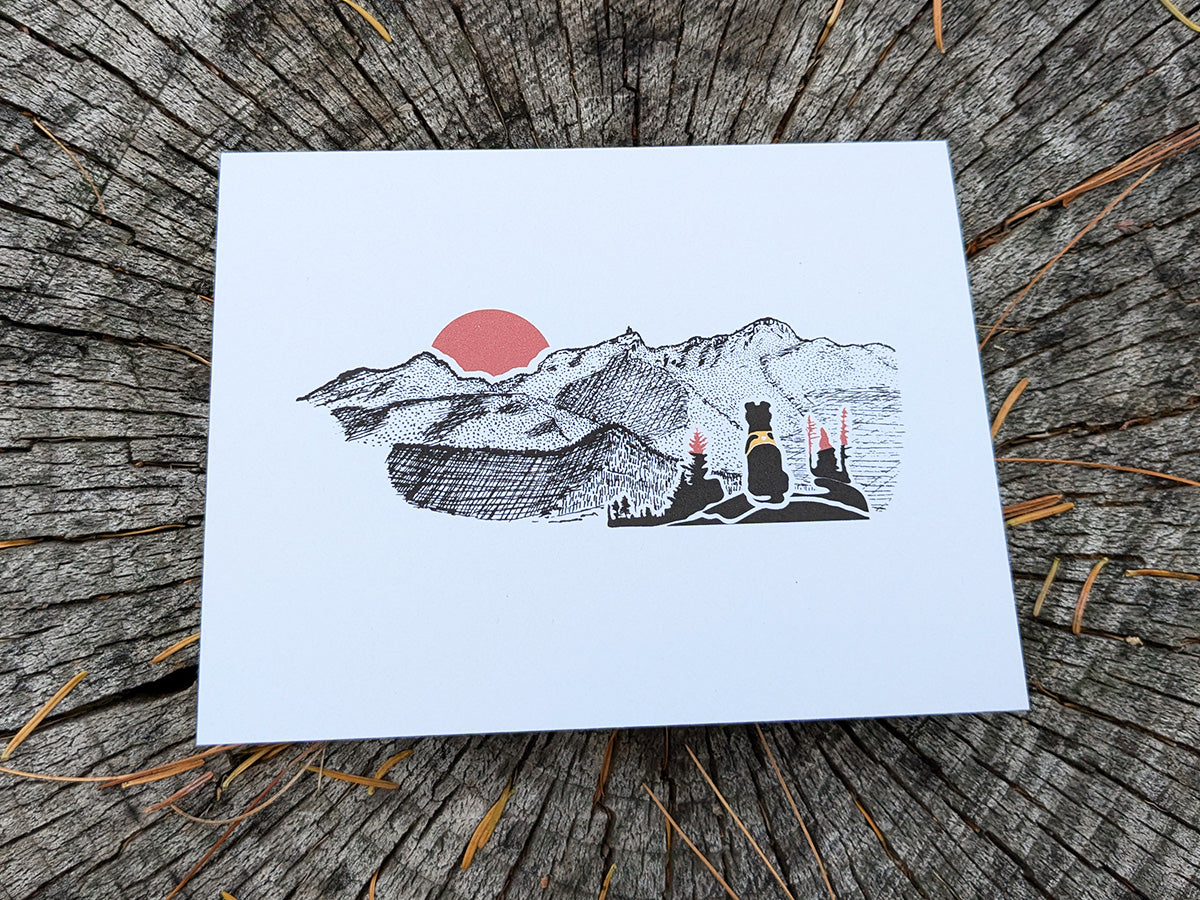 Greeting card with image of black dog looking at a red sunset behind mountains.