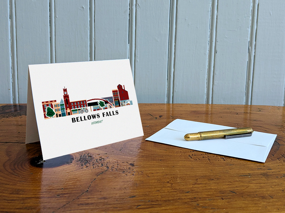 Greeting card with image of buildings arranged in a city skyline with the text Bellows Falls Vermont on a table with gold pen and envelope.