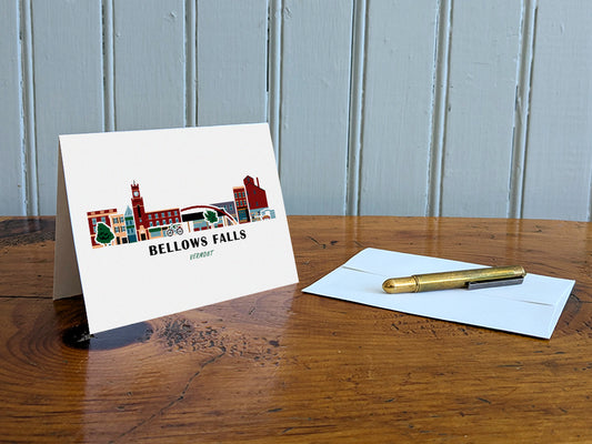 Greeting card with image of buildings arranged in a city skyline with the text Bellows Falls Vermont on a table with gold pen and envelope.