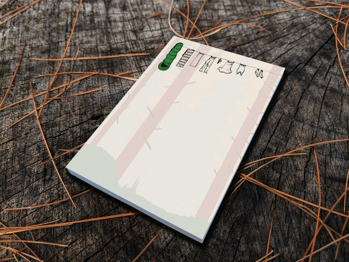 Notepad with illustration of clothes and sleeping bag hanging on a line in between pine trees in a forest, placed on wooden surface.