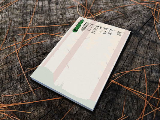 Notepad with illustration of clothes and sleeping bag hanging on a line in between pine trees in a forest, placed on wooden surface.