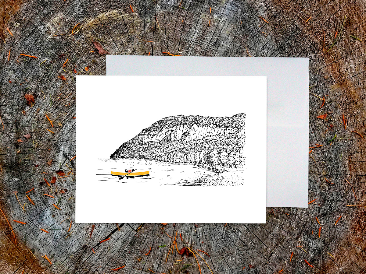 Greeting card featuring a dog paddling a yellow canoe on a lake with an ink illustrated mountain in the background, with a white envelope behind it and placed on a wooden surface.