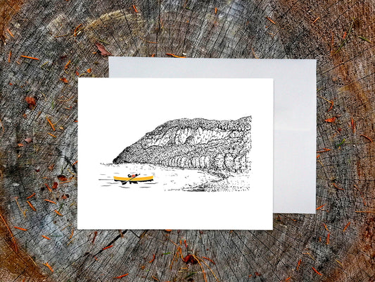 Greeting card featuring a dog paddling a yellow canoe on a lake with an ink illustrated mountain in the background, with a white envelope behind it and placed on a wooden surface.