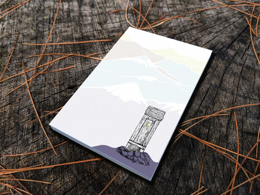 Notepad with an illustration of an outhouse in the mountains, placed on a wood surface.