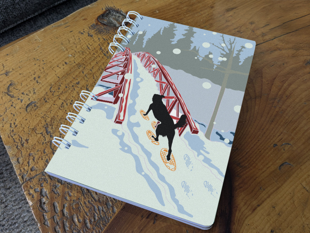A spiral-bound notebook with an illustration of a dog snowshoeing over a bridge, shown on a wood surface.