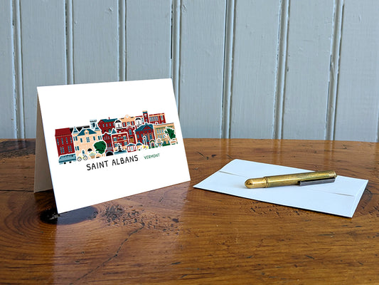 Greeting card with image of buildings arranged in a city skyline with the text Saint Albans Vermont on a table with gold pen and envelope.