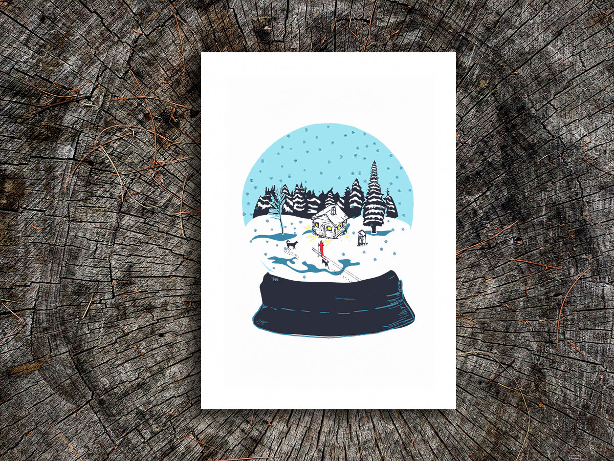 An art print featuring a snow globe illustration with a cabin, skier, and two dogs among a snowy landscape, displayed on a wooden surface.