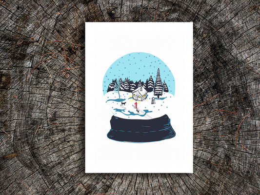 An art print featuring a snow globe illustration with a cabin, skier, and two dogs among a snowy landscape, displayed on a wooden surface.