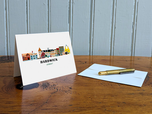 Greeting card with image of buildings arranged in a city skyline with the text Hardwick Vermont on a table with gold pen and envelope.