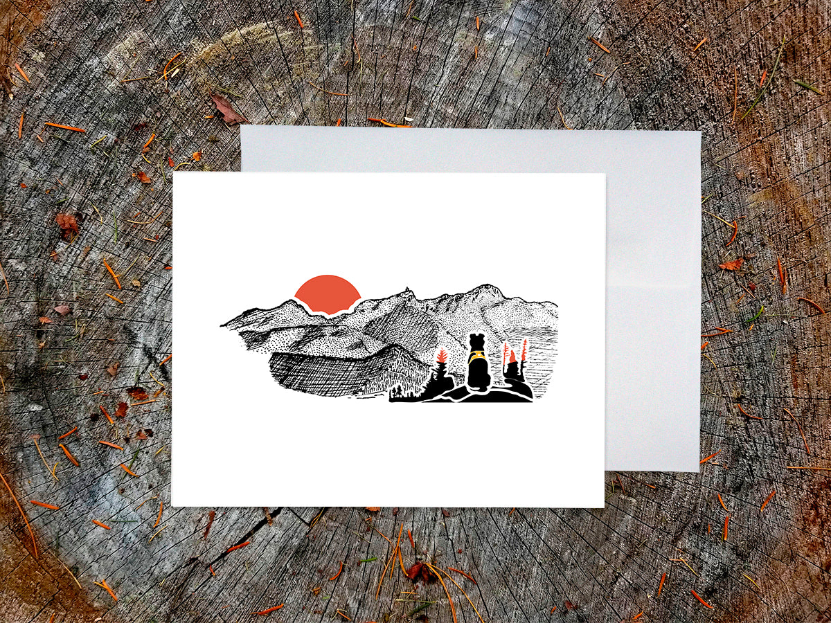 Greeting card with image of black dog looking at a red sunset behind ink illustrated mountains with envelope behind placed on a wood surface.