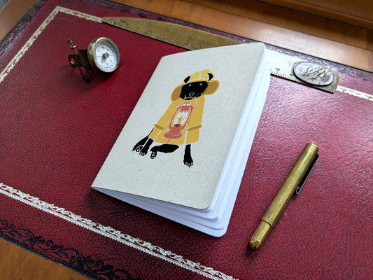 Notebook with dog in yellow jacket holding a lantern illustration on a red surface with a pen and small compass.