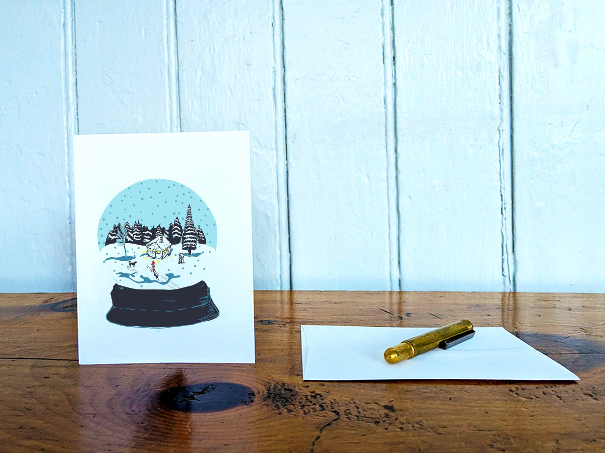 A greeting card with a snow globe illustration with a cabin, skier, and two dogs among a snowy landscape, displayed on a wooden table with an envelope and gold pen.