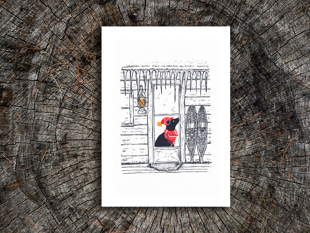 Art print featuring an illustration of a dog with red scarf and hat in a cabin covered in icicles, displayed on a wooden surface.