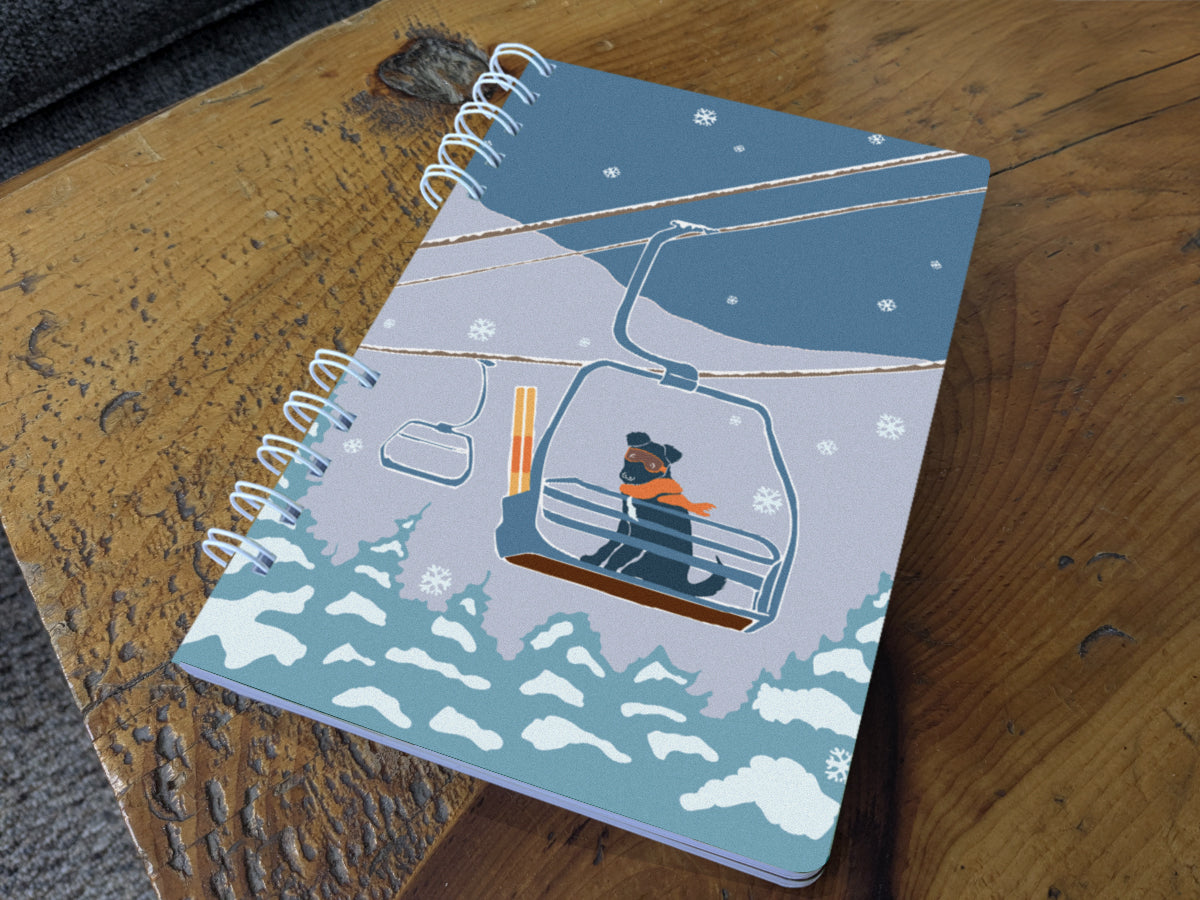 A spiral-bound notebook with an illustration of a dog riding a ski lift, on a wood surface.