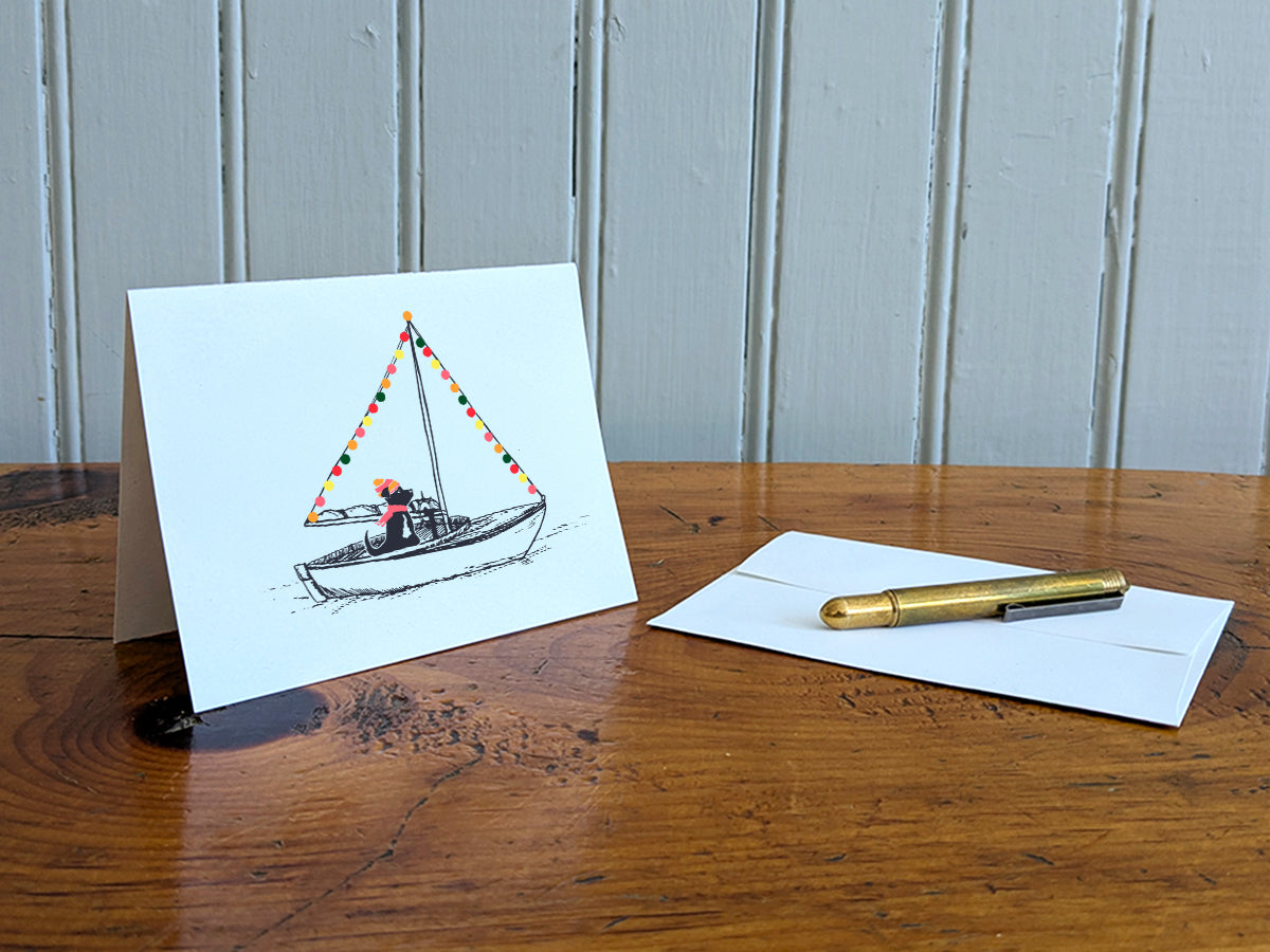 Greeting card with illustration of a black dog in a sailboat decorated with colorful lights, displayed on a wood table with white envelope and gold pen.