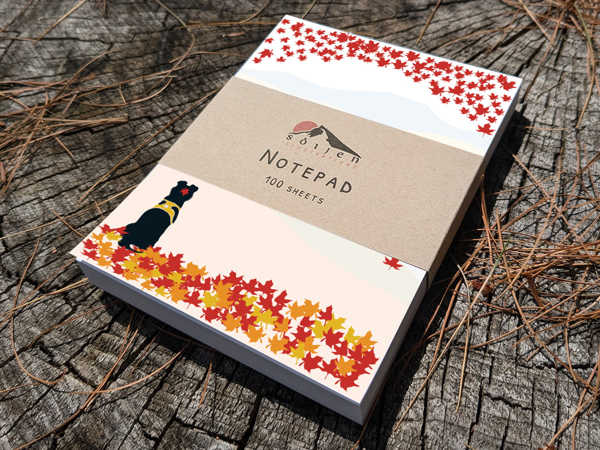 Notepad with a black dog among fall foliage on wooden background.