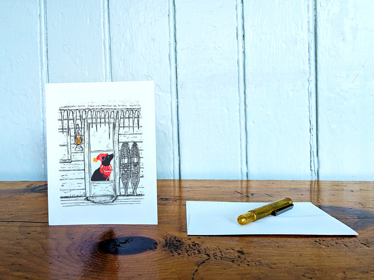 Greeting card with dog with red scarf and hat in a cabin covered in icicles, on a wooden surface next to a white envelope and gold pen.