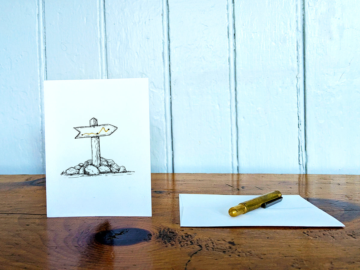 White card with a black ink drawing of a trail marker on a wooden surface with a pen and envelope.
