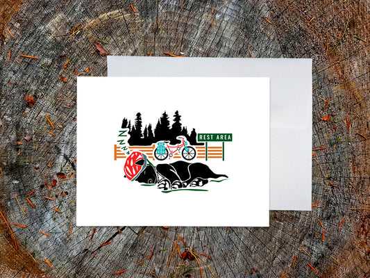 Greeting card of black dog with a red helmet sleeping with a bike and sign that reads Rest Area in the background, with a white envelope and placed on a wooden surface.