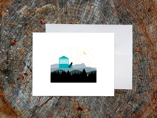 Greeting card of black dog sitting in a fire tower overlooking the mountains and stars, with a white envelope in the background and placed on a wooden surface.