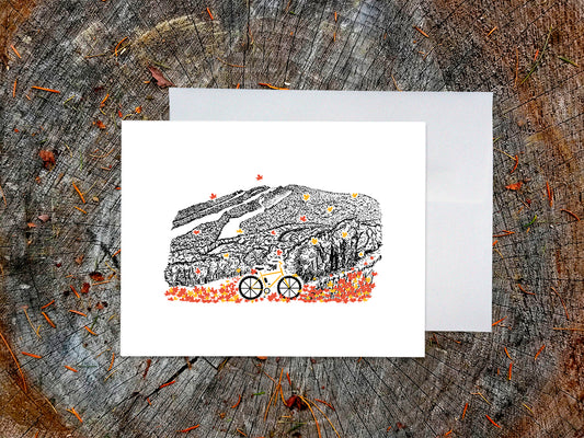 Greeting card with image of a mountain bike surrounded by brightly colored leaves with an ink illustrated mountain in the background, with an envelope behind it and placed on a wooden surface.