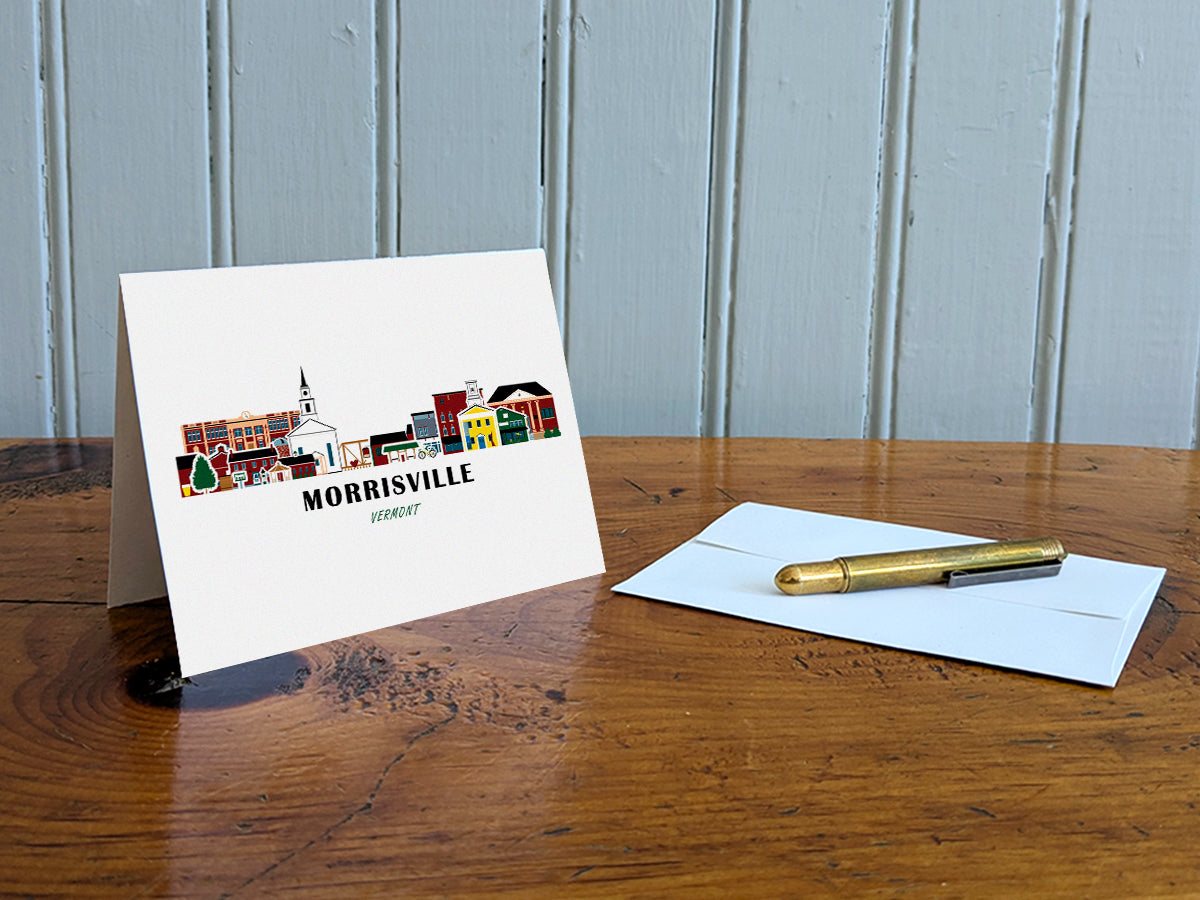 Greeting card with image of buildings arranged in a city skyline with the text Morrisville Vermont on a table with gold pen and envelope.