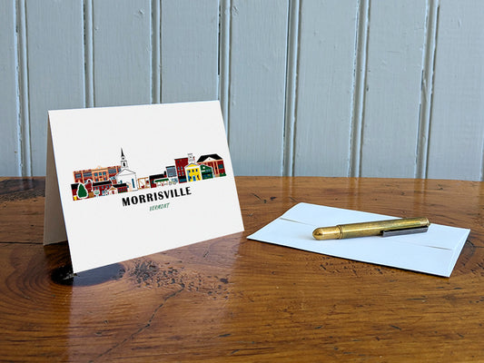 Greeting card with image of buildings arranged in a city skyline with the text Morrisville Vermont on a table with gold pen and envelope.