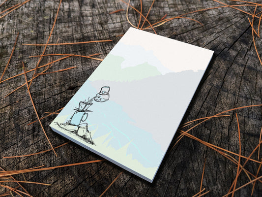 Notepad with illustration of pour-over coffee on a riverbank, placed on a wood surface.