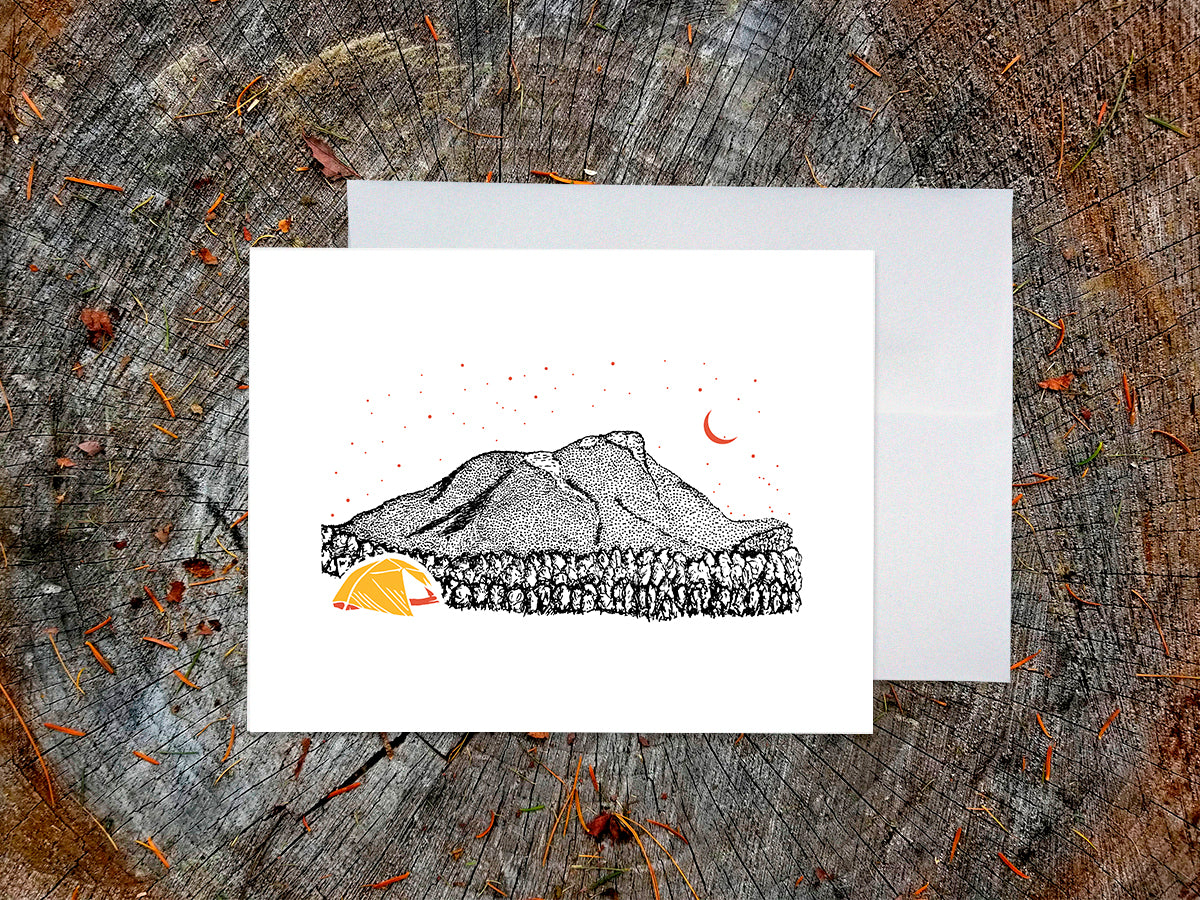 Greeting card with an image of an ink illustrated mountain and bright yellow tent under a starry sky, with a white envelope in the background and placed on a wooden surface.