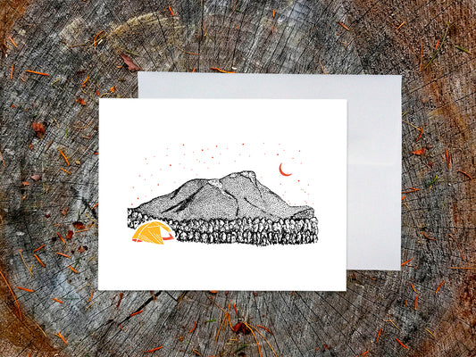 Greeting card with an image of an ink illustrated mountain and bright yellow tent under a starry sky, with a white envelope in the background and placed on a wooden surface.