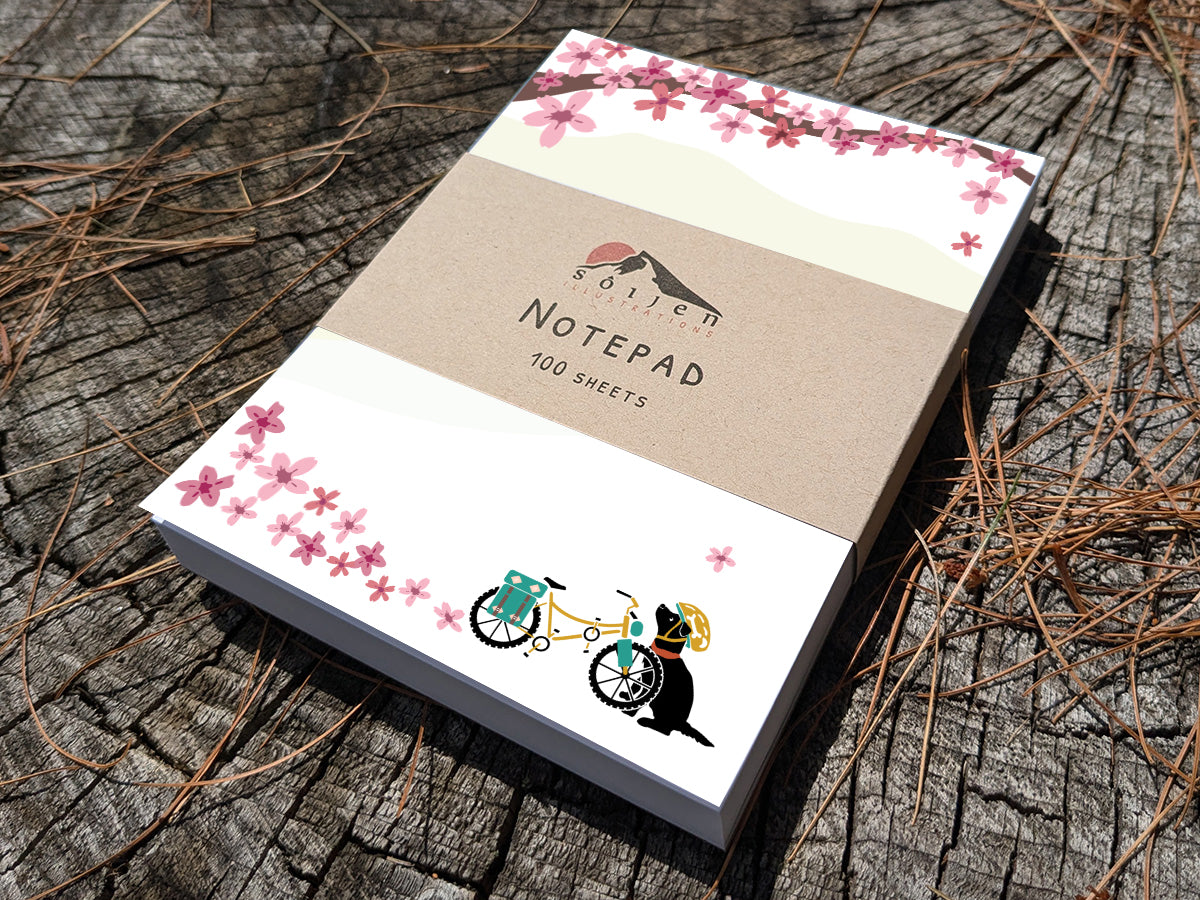 Notepad with a black dog next to bicycle among cherry blossoms on wooden background.