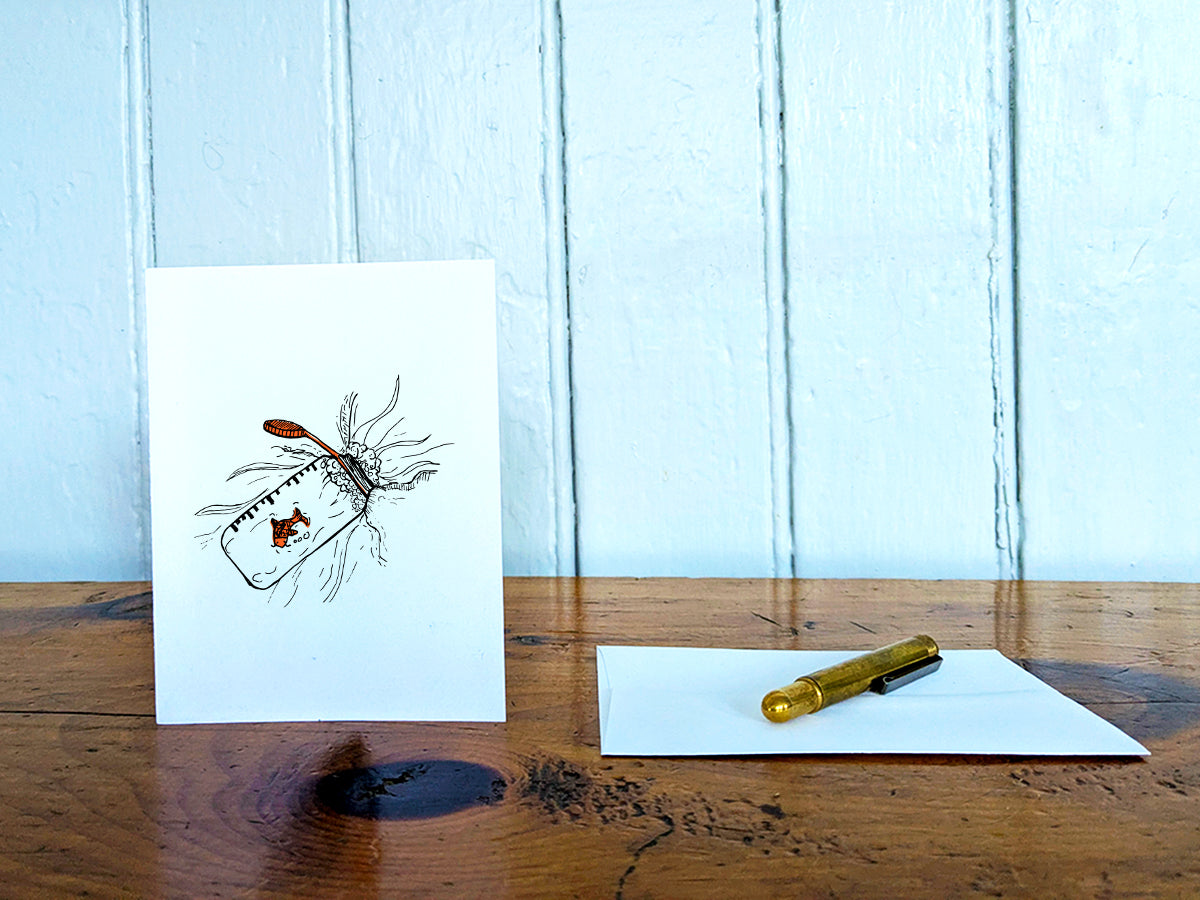 White card with a black ink drawing of a water bottle with red fish on a wooden surface with a pen and envelope.