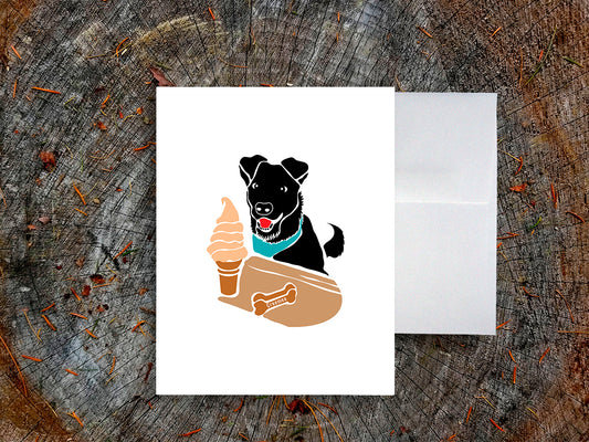 Greeting card with image of black dog looking at a soft serve ice cream on a table with a dog treat that says "creemee", with a white envelope in the background and placed on a wood surface.
