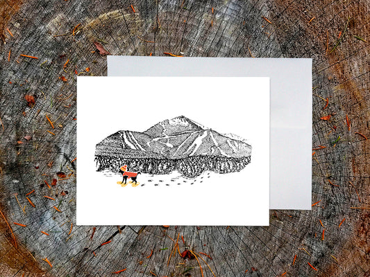 Greeting card with image of an ink illustrated mountain with ski trails and dog wearing a winter hat, vest and snowshoes, with a white envelope in the background and placed on a wooden surface.
