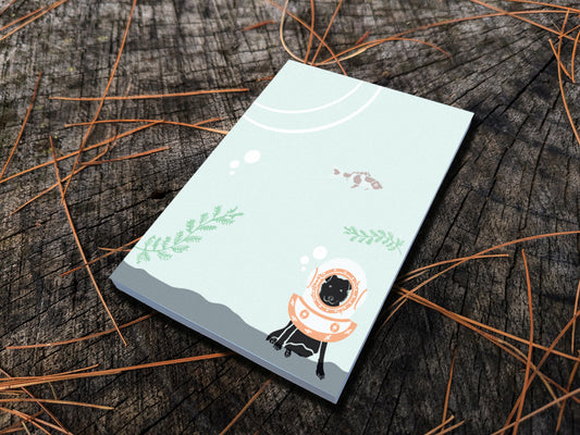 Notepad with a dog underwater with old school diving helmet and red fish, placed on a wood surface.