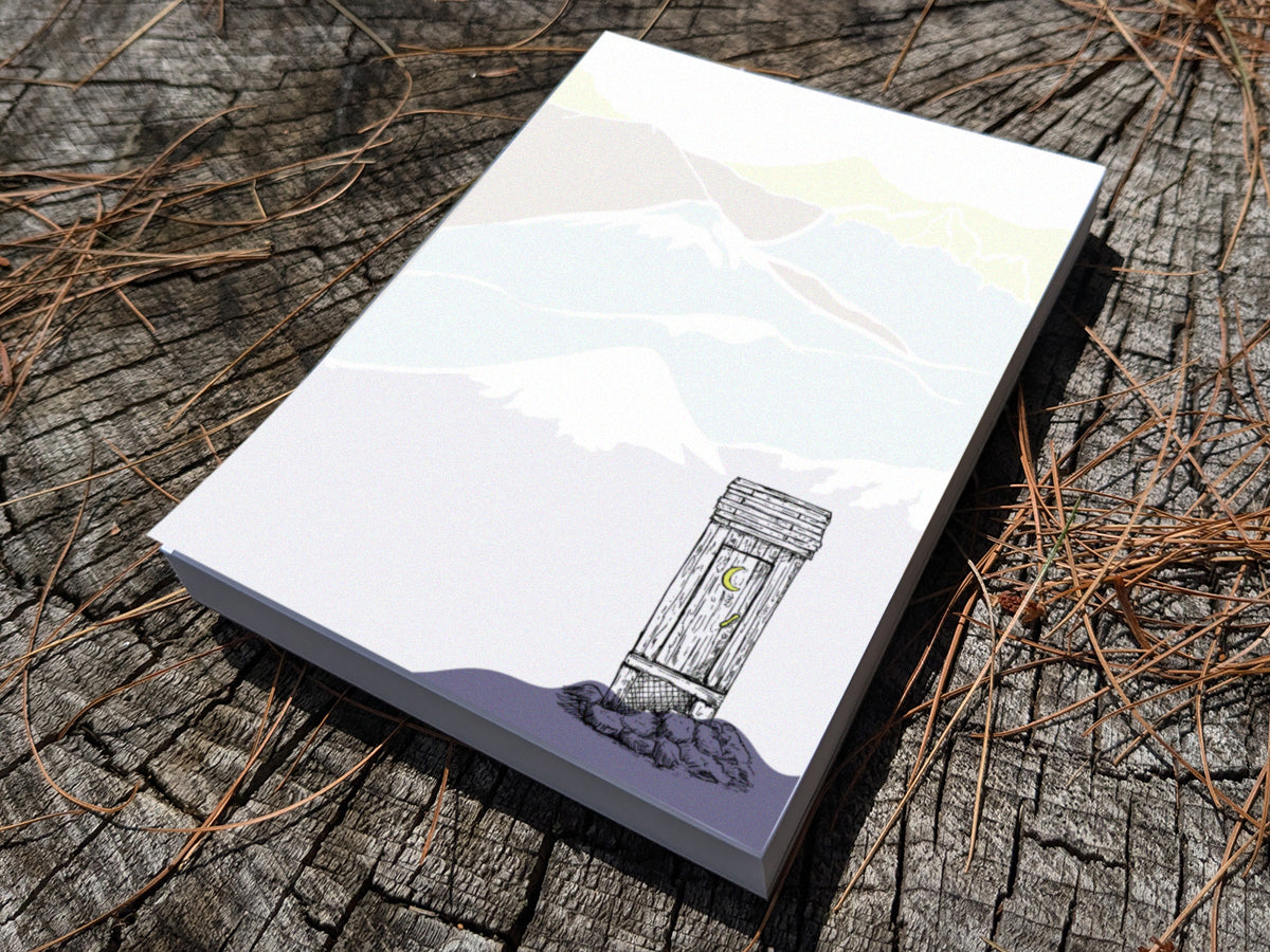 Notepad with an illustration of an outhouse in the mountains on a wood surface.