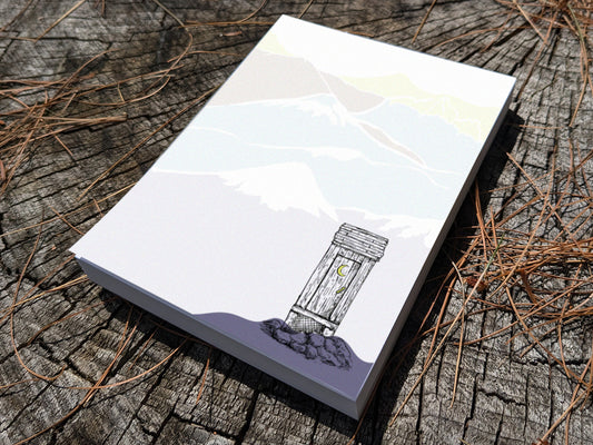 Notepad with an illustration of an outhouse in the mountains on a wood surface.