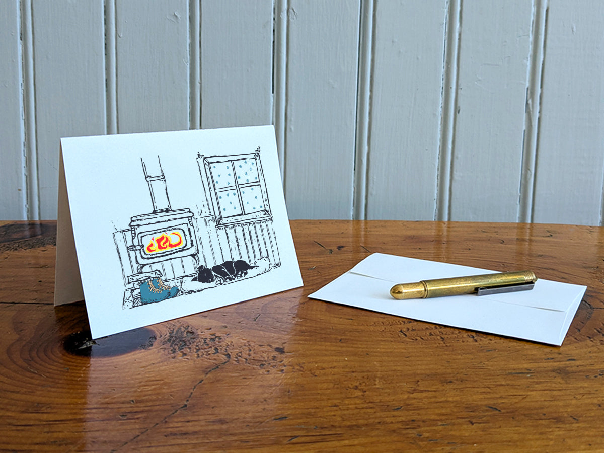A greeting card with a illustrated scene featuring a wood-burning stove, snow covered window, sleeping dog, and hiking boots, displayed on a wooden table with a white envelope and gold pen.