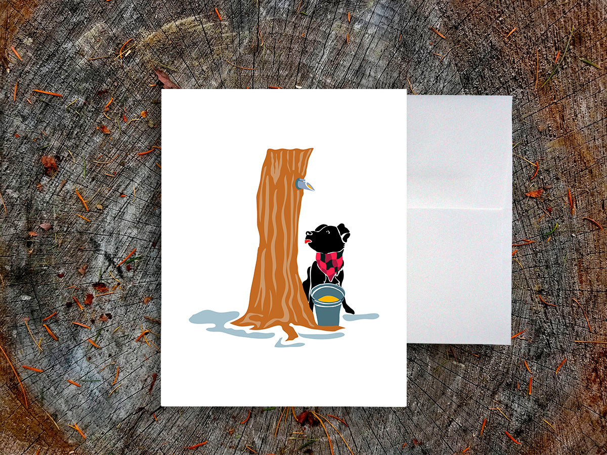 Greeting card with image of dog next to a maple syrup tap on a tree and bucket of maple syrup, with a white envelope in the background and placed on a wooden surface.
