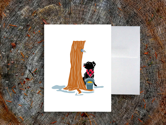 Greeting card with image of dog next to a maple syrup tap on a tree and bucket of maple syrup, with a white envelope in the background and placed on a wooden surface.