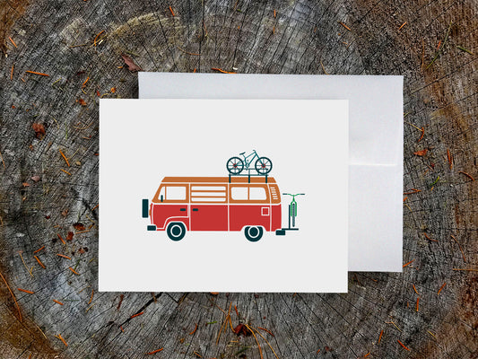 Greeting card with a red and orange camper van carrying two bikes with wood background