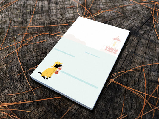 Notepad with an illustration of a dog in yellow hat and jacket holding a red lantern with a lighthouse in the background, placed on a wood surface.