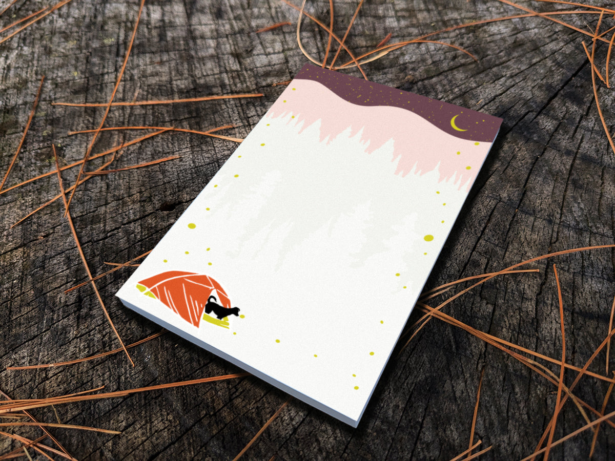 Notepad with an illustration of a black dog in red tent camping under night sky with fireflies, placed on a wood surface.