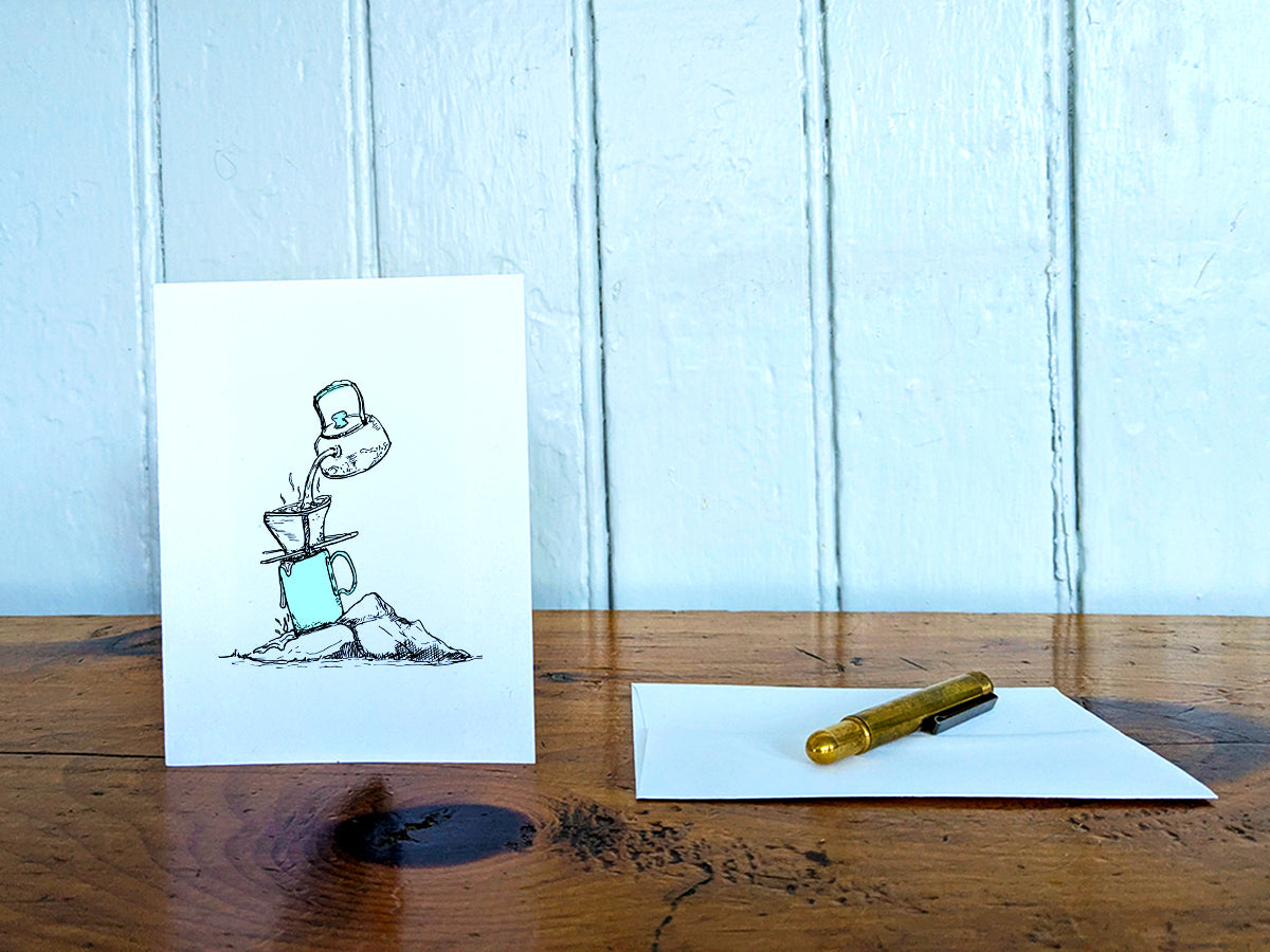 White card with a black ink drawing of pour-over coffee on a wooden surface with a pen and envelope.