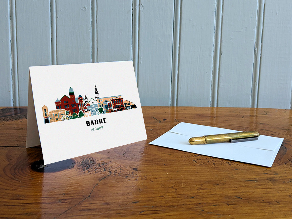 Greeting card with image of buildings arranged in a city skyline with the text Barre Vermont on a table with gold pen and envelope.