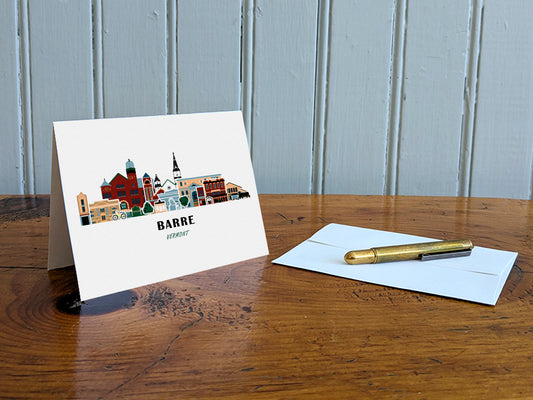 Greeting card with image of buildings arranged in a city skyline with the text Barre Vermont on a table with gold pen and envelope.