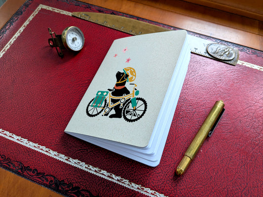 Notebook with dog with a bicycle illustration on a red surface with a pen and small compass.