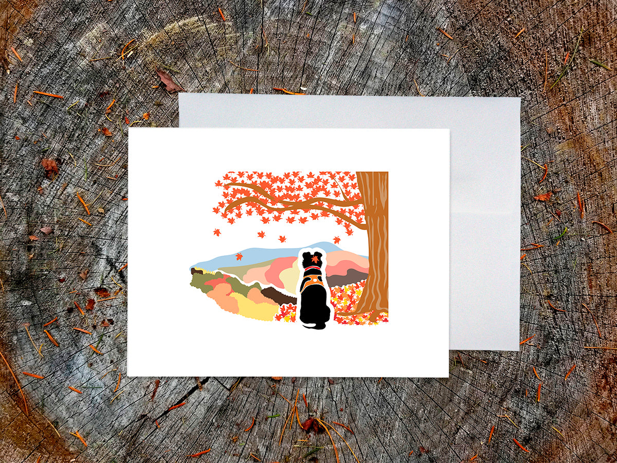 Greeting card with image of black dog underneath a maple tree with red fall foliage and mountains in the background, with a white envelope and placed on a wooden surface.