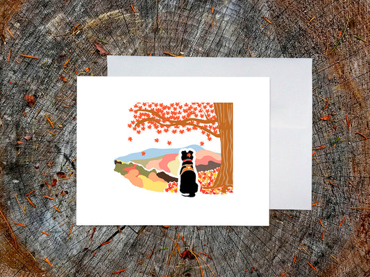 Greeting card with image of black dog underneath a maple tree with red fall foliage and mountains in the background, with a white envelope and placed on a wooden surface.