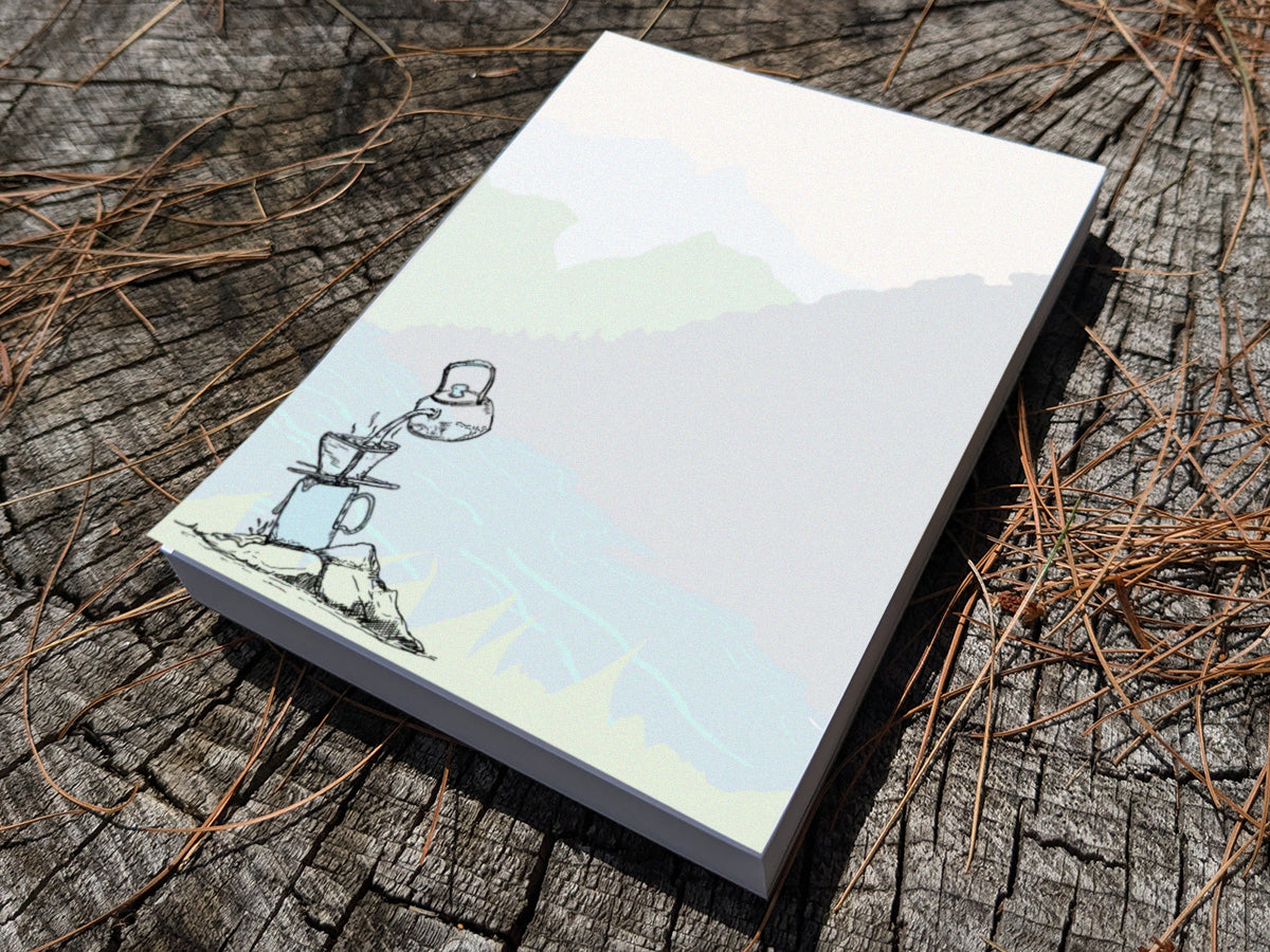 Notepad with illustration of pour-over coffee on a riverbank on a wood surface.